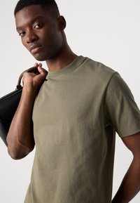 Olive green cotton T-shirt with a crew neckline, short sleeves, and a relaxed fit. The model carries a black leather bag with a handle.
