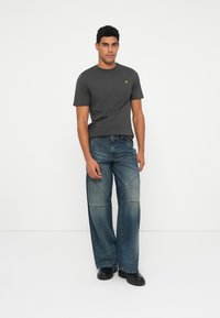 Homem a usar uma t-shirt de manga curta cinza escura, jeans azuis largos com detalhes costurados, e sapatos pretos, em pé contra um fundo branco simples.