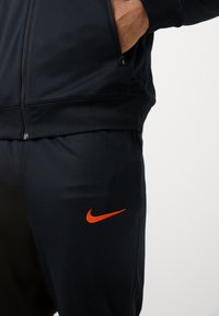 Nike sportos ruházat sötét navy színű anyagból, bal lábon narancssárga logóval, cipzáras zsebekkel és testhezálló kialakítással.