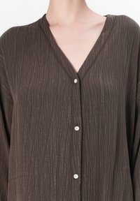 Gros plan sur une personne portant une chemise boutonnée à col en V de couleur marron foncé et texturée.