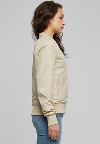 Bézs bomber dzseki sima felülettel, testhezálló mandzsettákkal és oldalzsebekkel, kék farmernadrág fölött viselve; oldalsó profilból bemutatva.