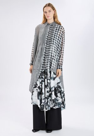 Donna in piedi che indossa una camicia lunga a strati con motivo a quadri bianco e nero sopra una gonna midi a fiori e pantaloni larghi neri.