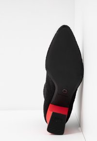 Zapato de tacón negro con parte superior de tela texturizada y puntera puntiaguda. Acento rojo en el tacón. Suela de goma negra lisa.