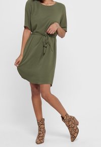 Oliivinvihreä, polvimittainen mekko, jossa on lyhyet hihat, vyö vyötäröllä, sileä kangas; yhdistetty ruskeisiin, käärmeennahkakuosisiiin nilkkabootseihin.