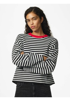 Femme aux longs cheveux bouclés porte un pull rayé noir et blanc avec un col rouge, bras croisés, sur un fond blanc uni.
