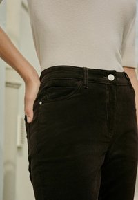 Juodos velveteen kelnės su siauru siluetu, su kontrastinga siūlėmis, priekinėmis kišenėmis ir mygtuko uždarymu. Medžiaga atrodo minkšta ir tekstūruota.