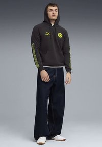 Felpa nera con cappuccio e accenti gialli, logo sul fronte e maniche patterned. Abbinata a jeans larghi blu scuro e sneakers bianche.