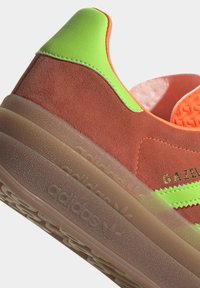 Sneakers Adidas Gazelle z brązową cholewką z zamszu, neonowozielonymi akcentami, pomarańczową wyściółką oraz przezroczystą gumową podeszwą z wytłoczonym logo.
