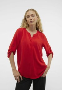 Blusa rossa a scollo a V realizzata in tessuto testurizzato con maniche a tre quarti arrotolate. L'orlo è leggermente curvato e la vestibilità è morbida.