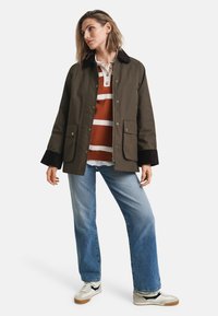 Veste en coton brun avec un col sombre, deux poches avant, portée sur un pull à rayures rouges et blanches, assortie avec un jean bleu et des baskets blanches.