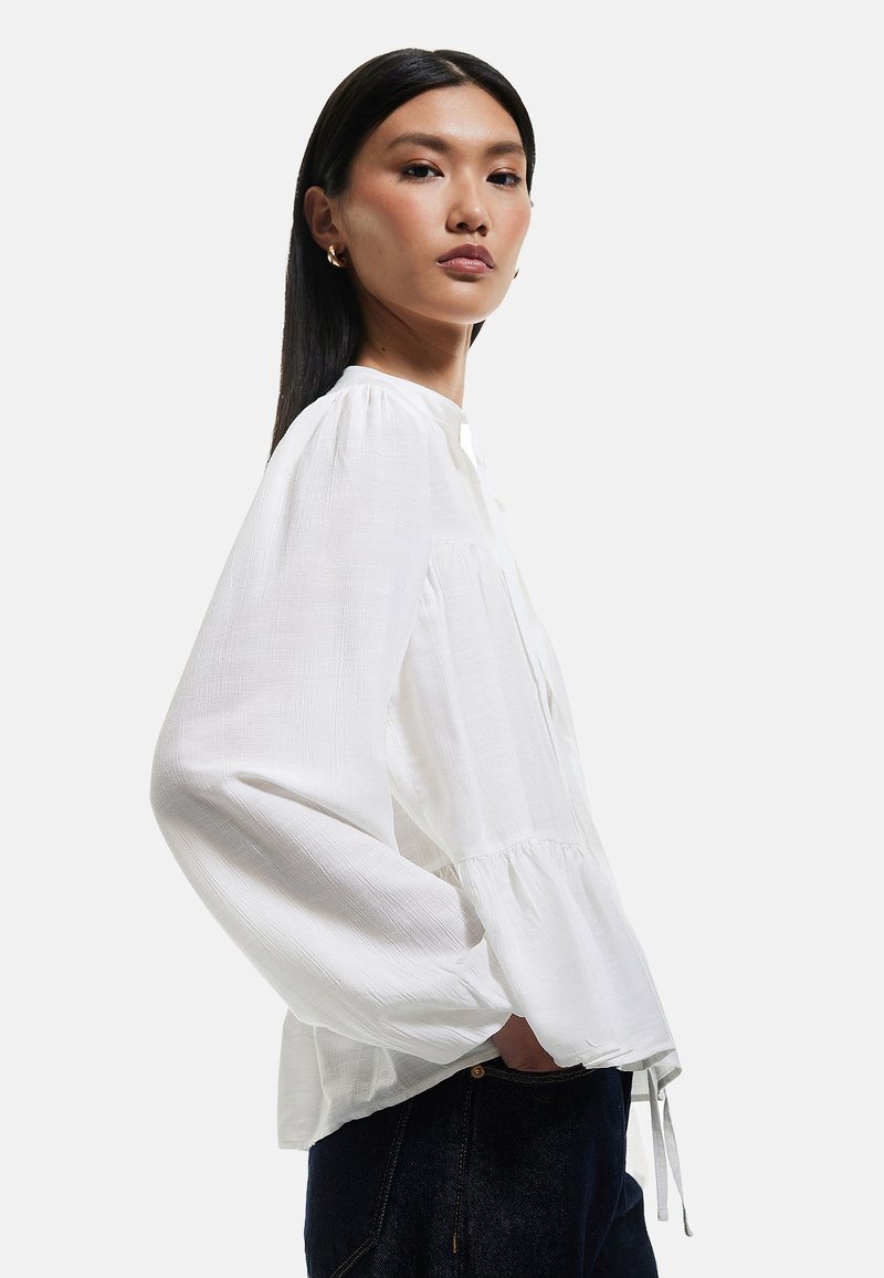 Blouse blanche à manches longues avec une finition texturée, un col rond et un ourlet ajustable par cordon. Présente des coutures d'épaule froncées et une fermeture à boutons.