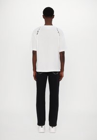 Chemise blanche à manches courtes avec des accents à motifs au dos, associée à un pantalon noir et des baskets blanches, vue de dos.