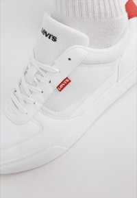 Valkoiset lenkkarit, joissa on verkkokangas ja keinonahkaa. Niissä on punainen Levi's-tagi, litteät nauhat ja kohokuvioitu kumipohja. Yksinkertainen muotoilu ilman kuvioita.
