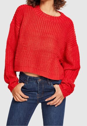 Žena má na sobě texturovaný, jasně červený krátký svetr s dlouhým rukávem a tmavě modré džíny, ruce má v kapsách.