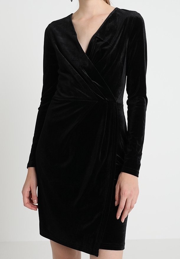 Robe portefeuille en velours noir à manches longues avec un col en V et un ourlet asymétrique portée par une personne debout.