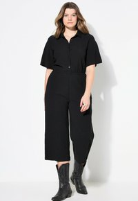 Mono negro de manga corta, con botones en la parte delantera, cinturilla elástica y piernas amplias y recortadas, combinado con botas vaqueras negras.