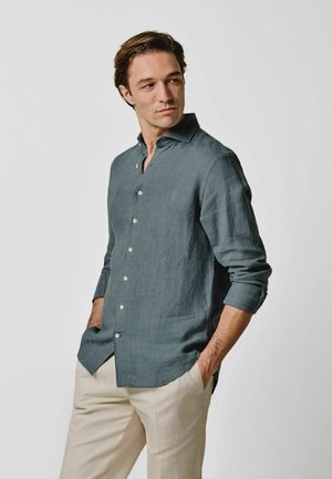 Mann trägt ein türkisfarbenes langärmliges Hemd mit Knöpfen und beige Hose, steht mit den Händen in den Taschen und schaut nach links.