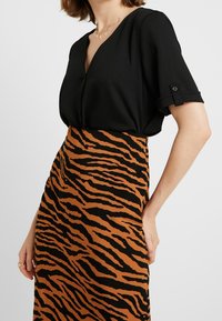 Kvinde iført en sort V-udskæringsbluse med op-rullede ærmer og en brun og sort tigermønstret højtaljet nederdel.