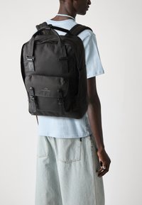 Sac à dos en tissu noir avec des bretelles réglables, un rabat avant et des fermetures à boucle. Design minimaliste avec un logo détaillé à l'avant.