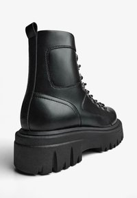 Ghete de gleznă din piele neagră, cu talpă supradimensionată și rotundă. Prevăzute cu ochiuri metalice și șireturi în față, cu o buclă pentru tragere la călcâi.