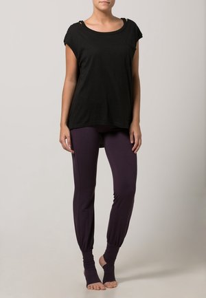 Kobieta ubrana w luźną czarną bluzkę z krótkim rękawem oraz dopasowane ciemnofioletowe legginsy z uchwytem na stopę, stojąca boso na jednolitym tle.