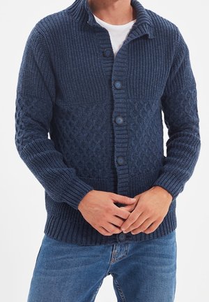 Man draagt een donkerblauwe gebreide cardigan met grote knopen over een wit shirt en blauwe spijkerbroek, handen samengevouwen ter hoogte van de taille.