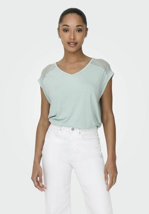 Licht mintgroene t-shirt met een v-hals, opgerolde mouwen en gemusterde gaasaccenten op de schouders, gedragen met witte high-waisted jeans.