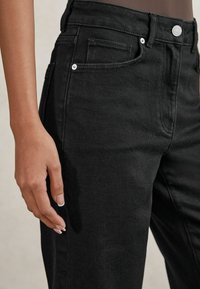 Los jeans de mezclilla negra presentan un diseño ajustado, cintura alta, detalles sutiles en los bolsillos y acentos de hardware metálico. Textura de tela suave visible.