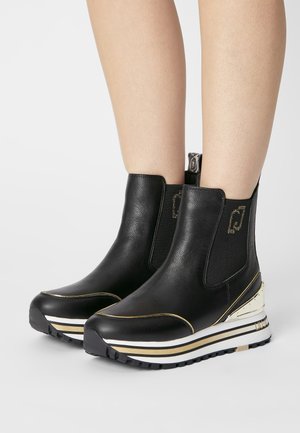 Chevilles et parties inférieures des jambes portant des bottines plateformes en cuir noir avec des accents dorés et des semelles blanches à rayures sur un fond uni.