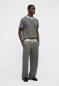 Μαύρος άνδρας που στέκεται με τα χέρια στις τσέπες, φοράει γκρι φανελάκι και φαρδιά παντελόνια φόρμας με λευκές λεπτομέρειες λογότυπου.