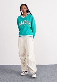 Τουρκουάζ φούτερ με λευκές και μαύρες ρίγες, με κείμενο "ALLESSE". Συνδυασμένο με κρέμ παντελόνι σε φαρδιά γραμμή και μαύρα-άσπρα παπούτσια.