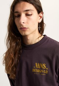 Bordowy sweter z okrągłym dekoltem, z złotym logo "VANS DESIGN CO. QUALITY GOODS". Model ma długie włosy i nosi srebrną kolczyk.