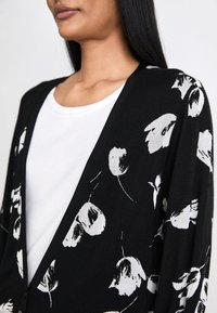 Svart kofta med vit blommönster, öppen framsidan design, mjukt tyg, rundad halsringning och texturerade detaljer på ärmar och nederkant.