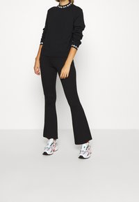 Sudadera negra con cuello y puños en contraste, combinada con pantalones negros acampanados. La modelo lleva zapatillas deportivas coloridas. Fondo blanco.