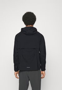 Mężczyzna z kręconymi włosami ubrany w czarną kurtkę z kapturem i ciemnoszare spodnie, pokazany od tyłu na jednolitym tle.