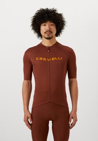 Korte mouw fietspak in bruin met contrasterend geel "castelli" logo. Voorzien van een ritssluiting aan de voorkant en een gepolsterd zitgedeelte voor comfort.