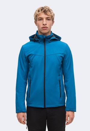 Jeune homme portant une veste à capuche bleue zippée avec des poches zippées noires, debout devant un fond uni.