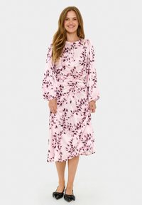 Rosa Kleid mit floralem Muster, langen Ärmeln, gerafftem Taillendetail und fließendem Stoff, der bis zur Mitte der Waden reicht, kombiniert mit schwarzen Schuhen.