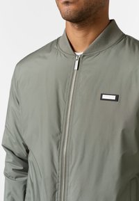 Giacca bomber verde oliva con zip frontale, colletto a coste e una piccola etichetta nera sul petto. Tessuto liscio e leggero.