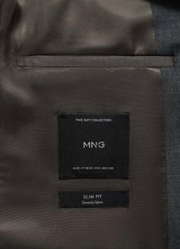 Črna tkaninska nalepka z belim besedilom "MNG" in "SLIM FIT." Material ima gladko teksturo s subtilnim sijajem, pritrjen na podlogo siva suknjiča.