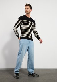 Suéter a rayas en negro y crema, textura acanalada, combinado con jeans de color azul claro. El calzado es negro, con un diseño deportivo y acentos grises.