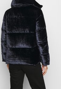 Giacca puffer di velluto blu scuro con colletto alto, cuciture orizzontali e una trama morbida e pelosa. Adatta per il clima fresco.