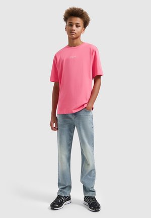 Tiener met krullend haar, gekleed in een roze T-shirt, lichtblauwe spijkerbroek en zwarte sneakers, staand tegen een effen achtergrond.