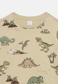 Μπεζ βαμβακερό t-shirt με πολύχρωμες εικόνες δεινοσαύρων και ηφαιστείου. Στρογγυλή λαιμόκοψη, κοντά μανίκια και ολόκληρο με παιχνιδιάρικο μοτίβο.