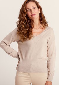 Beige Strickpullover mit V-Ausschnitt, strukturiertem Muster, langen Ärmeln und geripptem Saum. Weiches Material mit leicht glänzender Oberfläche.