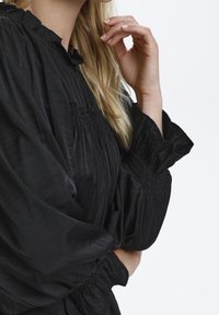 Svart blus med smockdetaljer, som har voluminösa ärmar och en rufsig halsringning. Tyget har ett mjukt, strukturerat utseende.