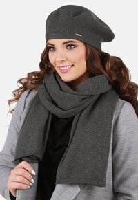 Berretto a maglia grigio e sciarpa coordinata, entrambi con una texture morbida. La sciarpa è avvolta attorno al collo con le estremità frangiate visibili.