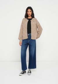 Beige stickad kofta med lös passform och strukturerat mönster, bärs över en svart skjorta. Kombineras med vida blå jeans och svarta sneakers.