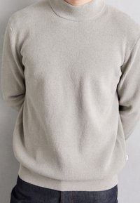 Pull gris clair côtelé avec un col montant, tissu texturé et coupe décontractée. Associé à un pantalon en jean foncé.