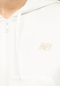 Κοντινό πλάνο ενός ατόμου που φοράει λευκό φούτερ με φερμουάρ και μπεζ λογότυπο New Balance στο στήθος και λευκά κορδόνια.